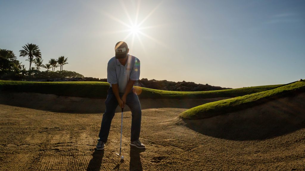 Pro Garratt Okamura chipping in a sand trap of golf course with sun behind