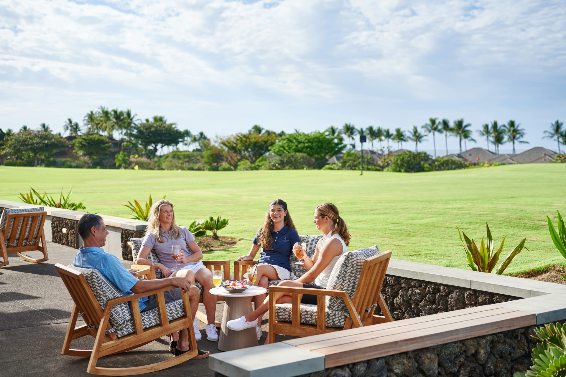 A family of four sits around a table in comfortable lounge chairs with a golf course in the background.