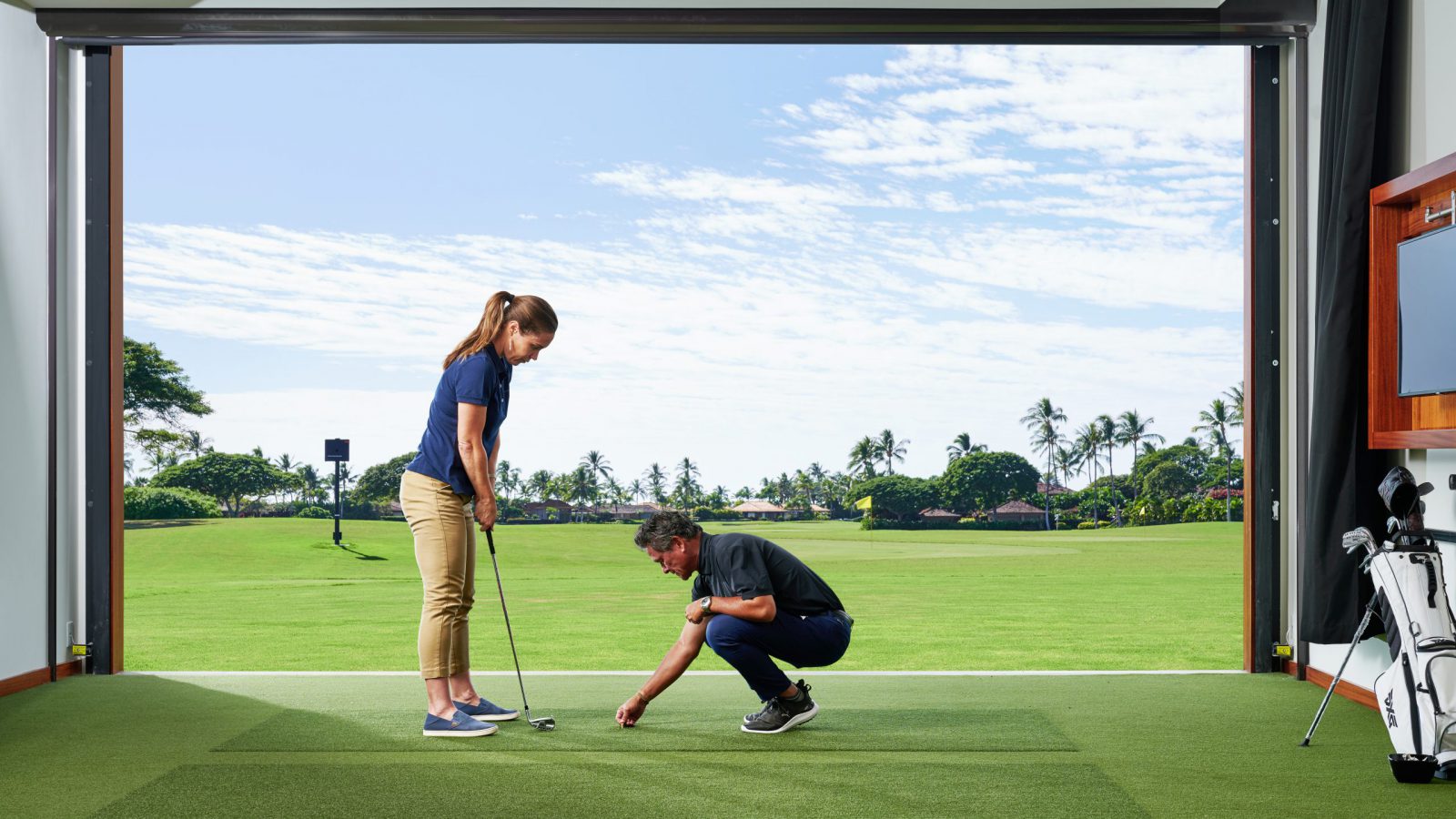An instructor places a tee on the indoor golf turf while the person taking the lesson readies her swing.